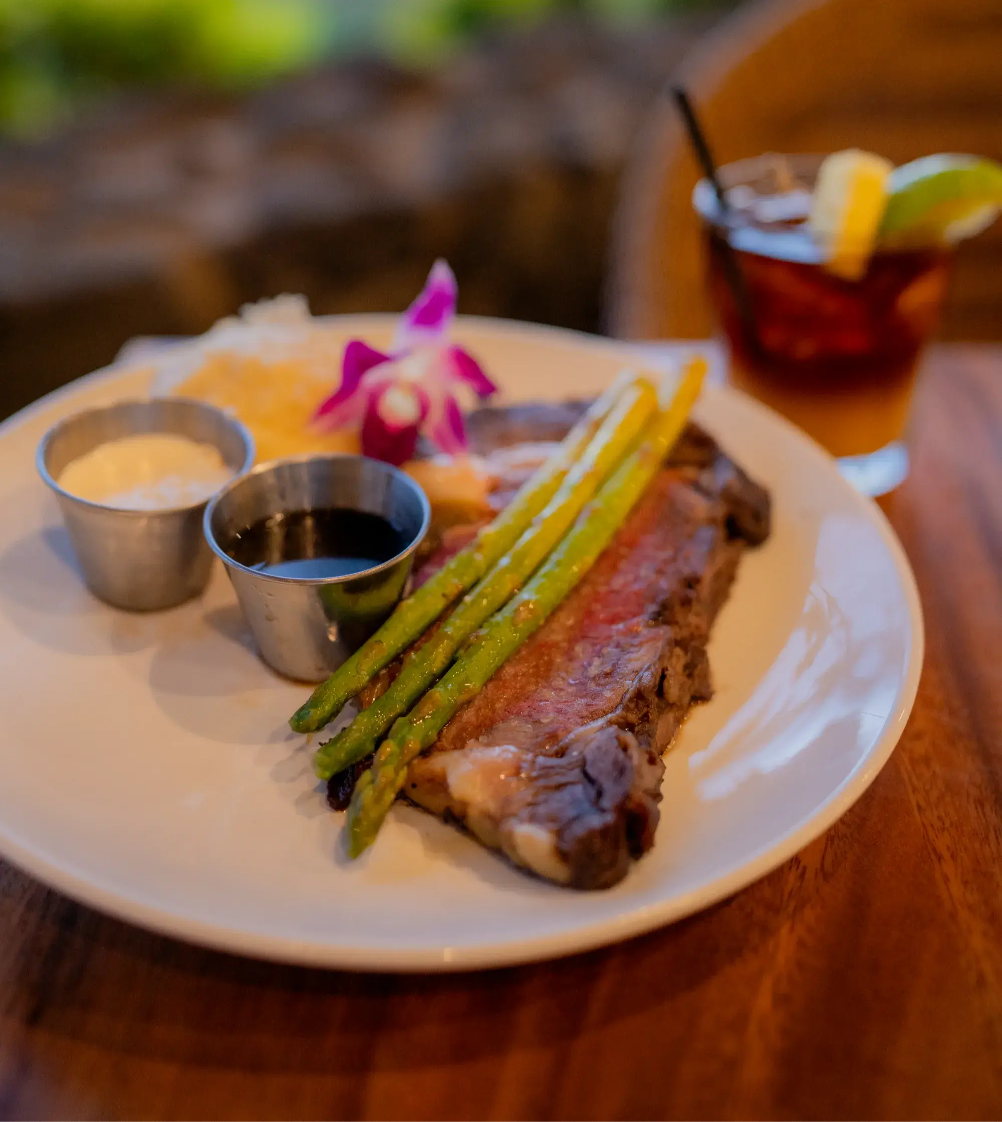 Kona Cut Prime Rib at Kona Inn Restaurant, sliced medium rare with grilled asparagus, au jus and horseradish cream, beside a cocktail