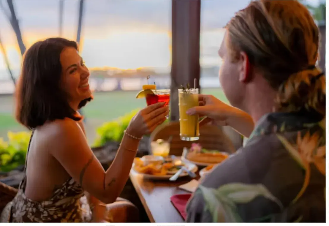 Two guests toasting cocktails at sunset on the open-air oceanfront lanai at Kona Inn Restaurant in Kailua-Kona