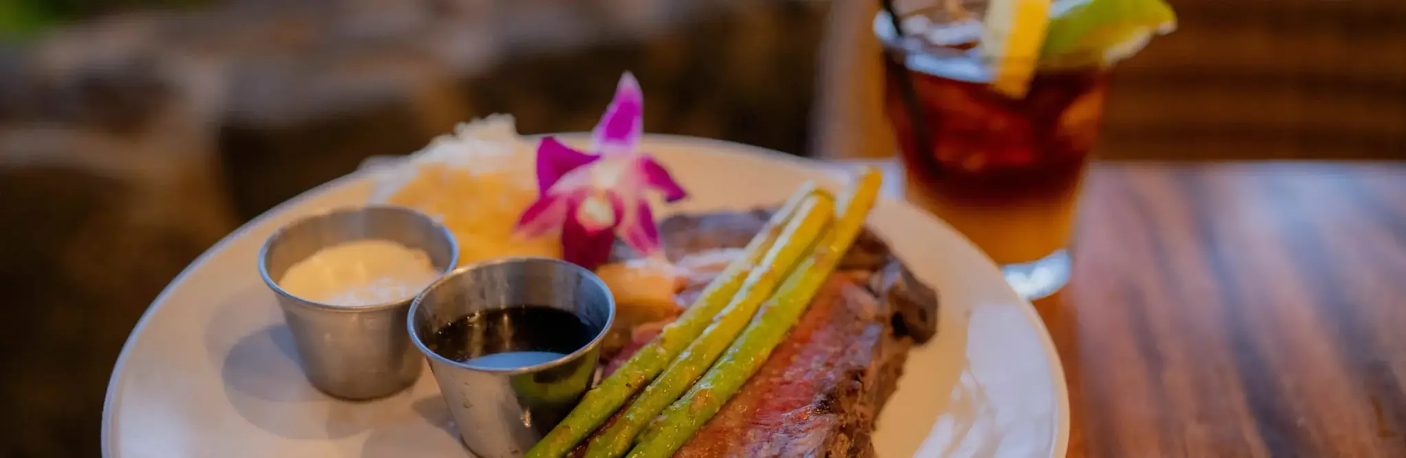 Kona Cut Prime Rib at Kona Inn Restaurant, sliced medium rare with grilled asparagus, au jus and horseradish cream, beside a cocktail on the oceanfront lanai