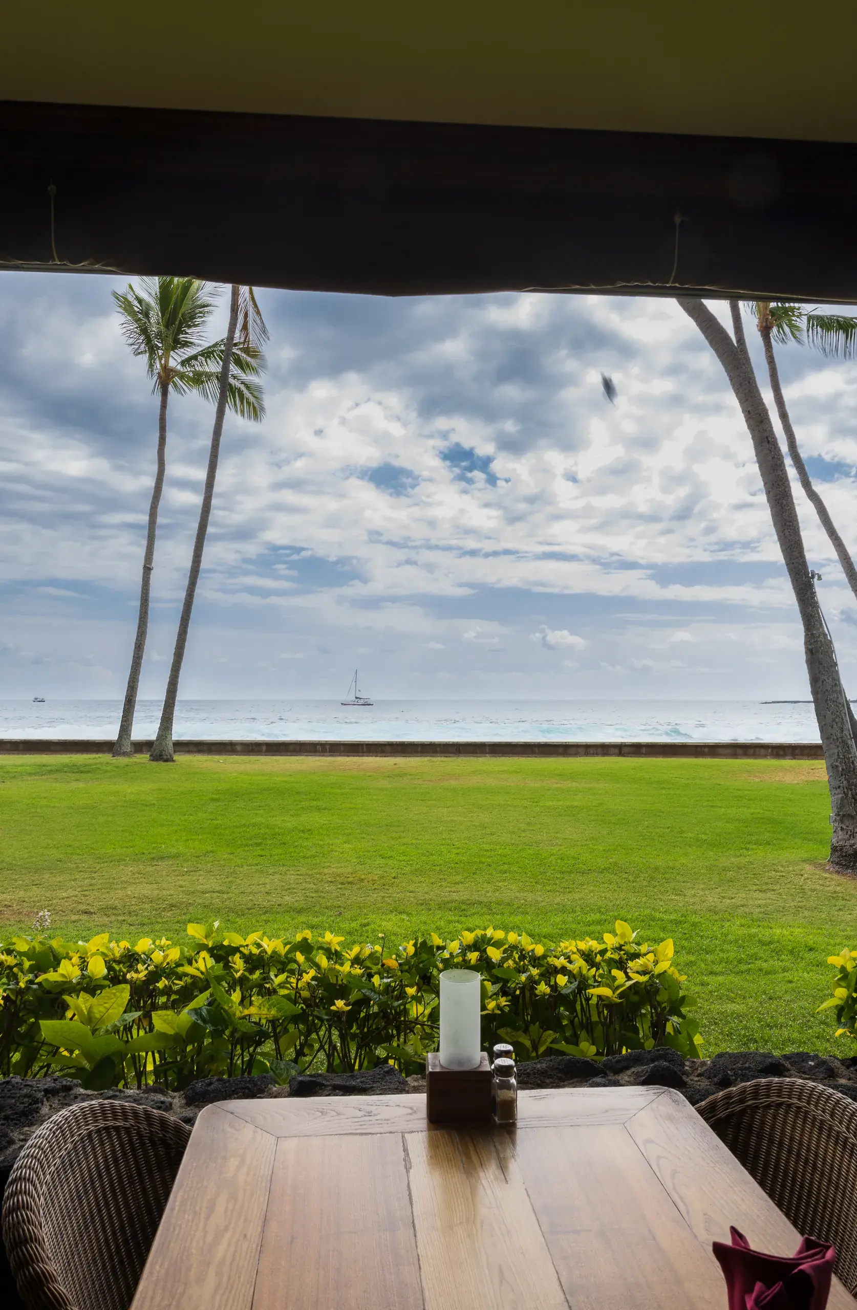 The view from a set table at Kona Inn Restaurant, looking past the lawn and palm trees to a sailboat on Kailua Bay